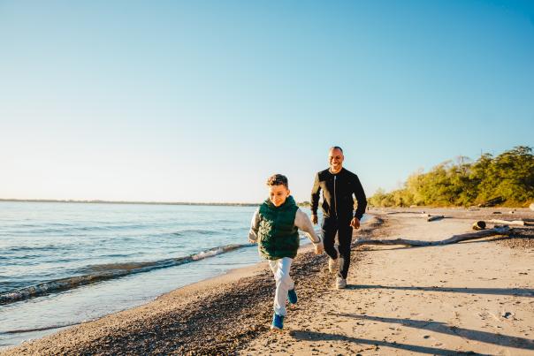 A child runs along a quiet sandy beach while an adult follows close behind, both smiling and enjoying the shoreline on a sunny autumn day. Gentle waves lap the shore, with trees and driftwood lining the beach in the background.
