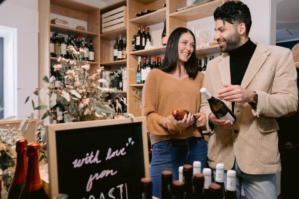 Smiling couple browsing wine bottles in Coast Wine House with wooden shelves filled with various wines and a floral display in the foreground, creating a warm and inviting atmosphere.