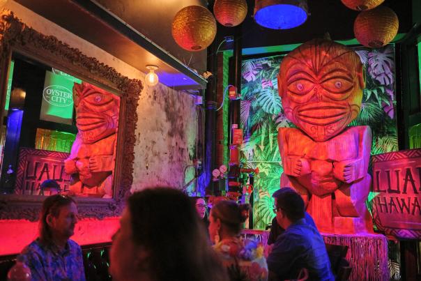 A vibrant bar scene featuring a large wooden tiki statue surrounded by colorful lighting and tropical decor. Guests are seated at tables, enjoying the atmosphere.