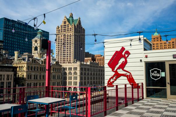 Red and white rooftop bar called The Aviary with patio seating, string lights, and a large red silhouette mural, set against the Milwaukee skyline.