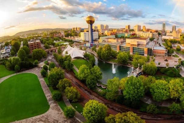 A wide-angle, panoramic aerial view of World’s Fair Park in Knoxville, Tennessee, at sunset, featuring the iconic Sunsphere, lush green landscaping, walking paths, and a central pond with a fountain, with the downtown city skyline in the background.