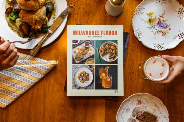 Milwaukee Flavor: The Cookbook rests on a wooden dining table surrounded by a meal. A hand holds a fork over a plate of chicken, vegetables, and potatoes on the left, while another hand holds a latte with heart-shaped foam art on the right. Around the book are a striped napkin, a floral plate, and a dessert plate with slices of cake.