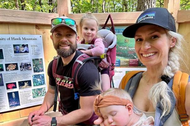 Ready for a spring hike, a young couple stands in front of informational signs at Hawk Mountain Sanctuary. The man wears a backpack holding his young daughter and the mother wears a harness that holds a sleeping little girl.