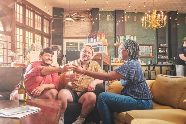 Three friends sit on couches laughing and toasting glasses of wine.