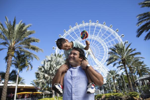 A boy on top of his dad's shoulders, while smiling and holding a lollipop, in front of The Wheel at ICON Park