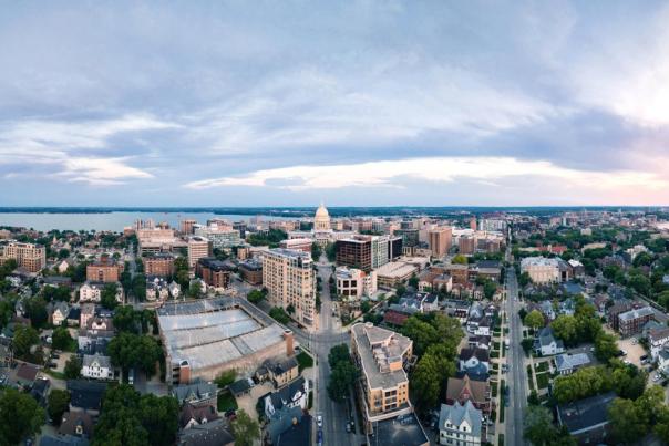 A panoramic view of a vibrant city skyline at sunset, with a lake in the background and tree-lined streets below. The scene captures a mix of modern and historic architecture.