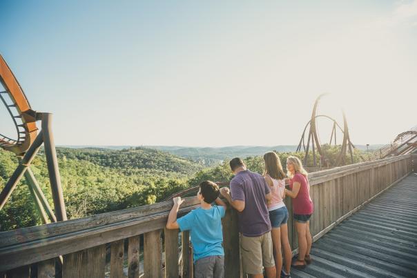 A wide, high-angle shot of a family of four looking out at the panoramic view from an elevated wooden boardwalk at Silver Dollar City in Branson, Missouri. On the left, a loop of an orange roller coaster track rises into the frame. In the center, a man in a purple shirt and khaki shorts stands with a young boy in a bright blue shirt to his left, and a young woman in a pink top and denim shorts to his right. Next to her stands another woman in a pink sleeveless shirt and shorts. All four have their backs to the camera, leaning against a rustic wooden railing to admire the vast expanse of rolling green Ozark hills stretching across the horizon under a clear sky. In the background on the right, the large, sweeping metal tracks and loops of the Wildfire roller coaster stand tall against the bright, sun-lit sky.