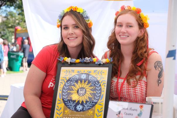 Two young smiling women with long hair are wearing traditional polish flower crowns while holdiing up Polish art at Muskegon Polish Festival