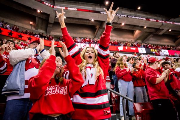 A white woman yells and raises her hands in the air in the crowd at a men's hockey game at the Kohl Center. She is wearing a red Badgers hockey jersey.