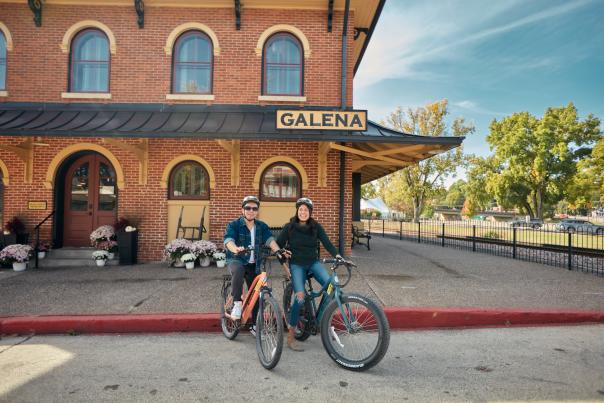 Two people on bicycles in front of the Visitors Center in Galena.