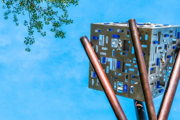 Steel sculpture titled city built on timber. 4 steel post cross each other to support steel box punctuated with squares, some are filled with blue and green colored glass. All set against blue sky with tree branch in upper left corner