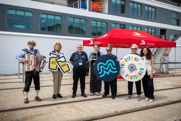 A welcoming committee with an accordion player stands before a cruise ship, holding Milwaukee-themed signs including an MKE logo and "City of Festivals" circular display, under a red Viking Cruises tent.