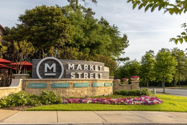 The entrance sign for Market Street is a rounded brick wall with two tiers. The top tier is gray brick bearing the name of the shopping center; the lower tier is light / beige brick with teal plaques bearing the names of various shops. The sign is surrounded by bright pink flowers and lush green trees.