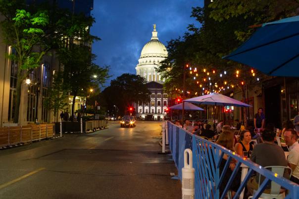 A lively street scene at night, featuring outdoor dining under string lights, with a prominent dome building in the background.