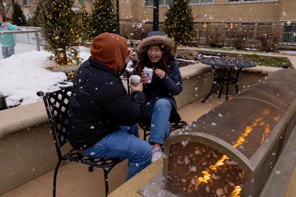 Two friends dressed in winter coats drinking coffee by an outdoor firepit while snow falls around them.