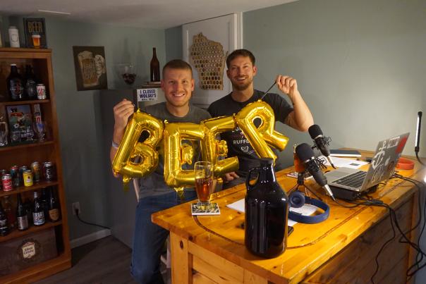Two white men hold shiny gold balloons that spell beer.