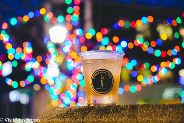 Clear plastic cup with round downtown Muskegon social district label sits on wall before strings of glowing holiday lights