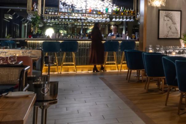 Tables and chairs in a fine dining restaurant leading to a brightly lit bar at the end of the room.