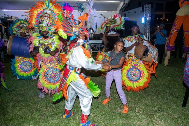 A group dressed in brightly-colored traditional Caribbean Festival attire dancing with a young girl and playing drums in front of an outdoor stage in Pompano Beach