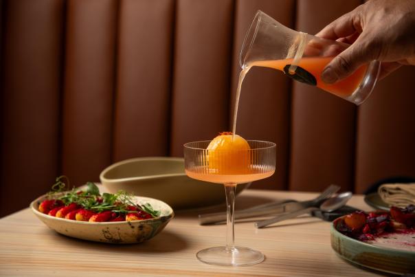A hand pours an orange liquid from a small carafe onto a frozen orange ball in a wide cocktail glass surrounded by trays of strawberries and other appetizers