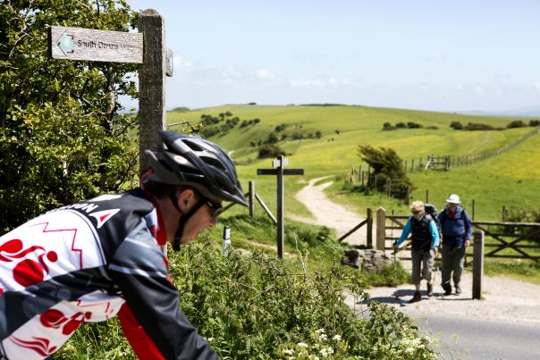 A mountain biker rides along the South Downs Way, with walkers on a trail in the background