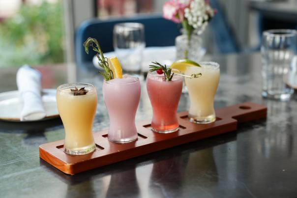 A flight of four frozen cocktails sit on a tray on the edge of a State Fare patio table. One is yellow with an anise seed pod floating on top; the second is pale purple with a lemon slice and herb sprig; the third is light red with a cranberry and sprig of rosemary; the last is pale yellow with a lime wedge.