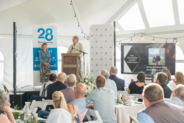 A speaker at a podium addresses an audience in a tented venue, with tables set for a formal event. A screen displays an award presentation.