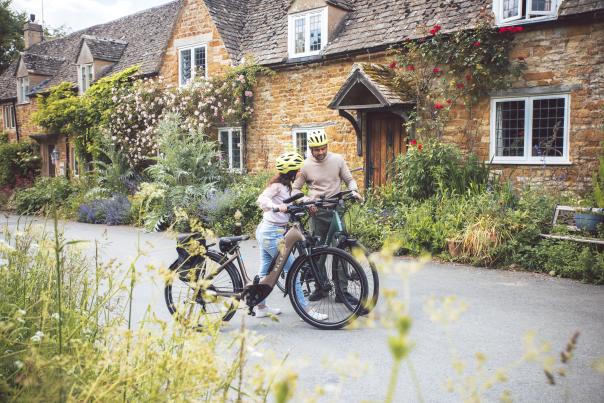 A couple have dismounted their bikes in front of a stunning cottage, with red roses climbing over it's porch, and are looking at their map
