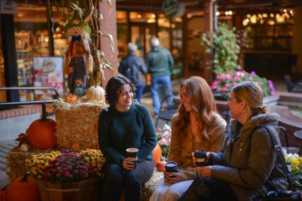 Three people sip coffee in the courtyard at the market with haybales and pumpkins.
