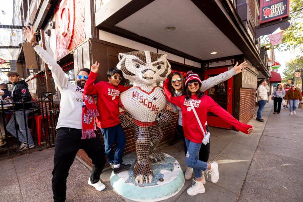 A family wearing UW Badger merch posing next to a Bucky the Badger statue outside of State Street Brats.