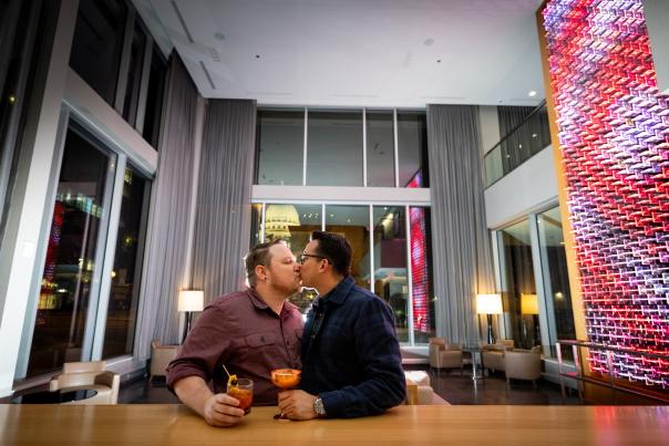 Two men at a hotel bar kiss with the Wisconsin State Capitol building lit up in the background.