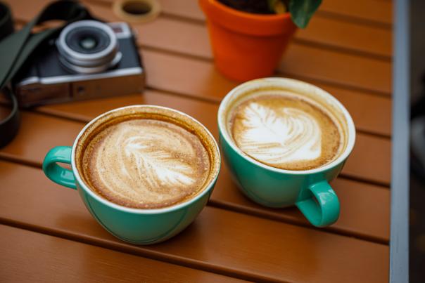 Two mugs with latte art on top, sitting on a wooden table, with a camera in the background.