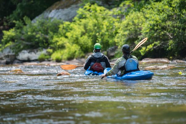 Two kayakers with blue boats and orange paddles paddle down a river with large boulders in the near distance.