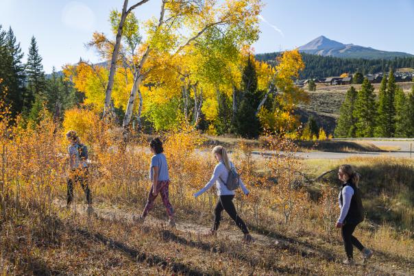 Four women walk along a trail surrounded by vibrant autumn foliage, with mountains and a clear sky in the background.