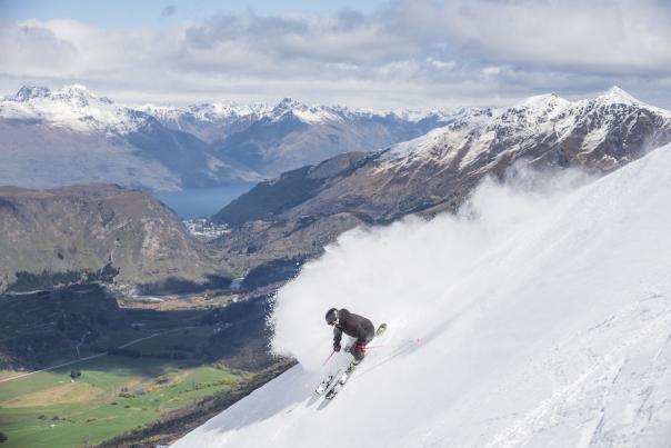 Person snowboarding down a mountain with views of green valley and mountains in the background