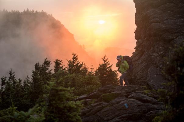 A orange sun is illuminated between two mountains rising on either side of a hiker walking a path between summits.