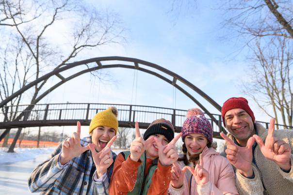 An adult man and three kids hold up their fingers in the shape of Ws while standing on a frozen lake with a bridge in the background