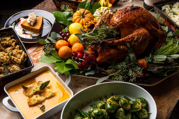 A Thanksgiving spread featuring a beautifully cooked turkey (top right) garnished with small oranges, lemons, and various types of miniature pumpkins. Dishes around the turkey (from left to right, top to bottom) include a slice of pumpkin pie, green bean casserole, a creamy orange soup, and roasted brussels sprouts.