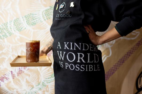 An iced coffee drink in a glass on a wooden tray held by a barista who's apron reads "a kinder world is possible"