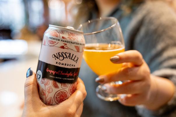 A close up of a can of NessAlla Komucha cheersing a glass full of the kombucha.