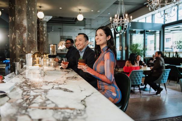 Two men and one woman sitting at a marble bar, enjoying colorful cocktails