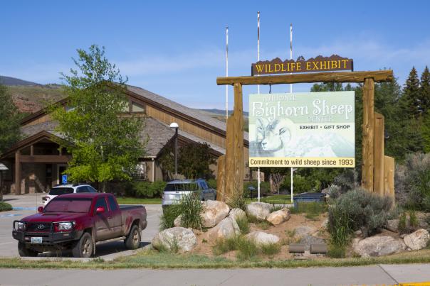 Exterior view of the National Bighorn Sheep Center in Dubois, Wyoming, featuring a large wooden sign in the foreground, a red pickup truck in the parking lot, and the center's rustic building under a clear blue sky