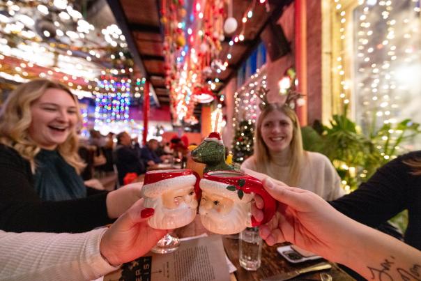 A group of women at a restaurant decked out in Christmas lights, cheersing their Santa mugs