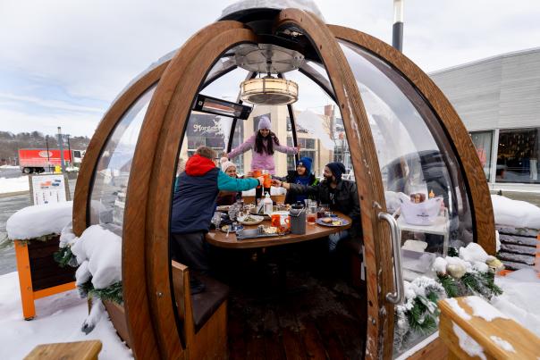 A group of friends cheersing their drinks while dining in an outdoor dome with snow around the outside of the dome.