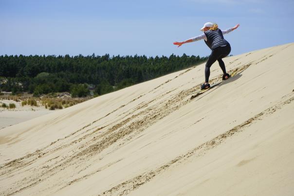 A person sandboards down a steep sand dune with trees in the distance