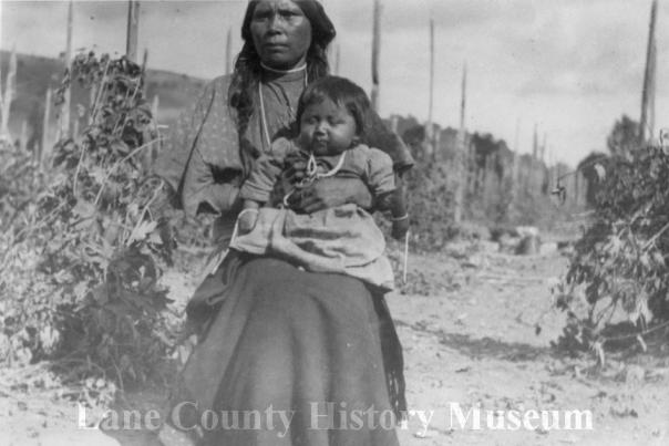 An Indigenous woman with a child sits in the middle of a hop farm in this black and white image.