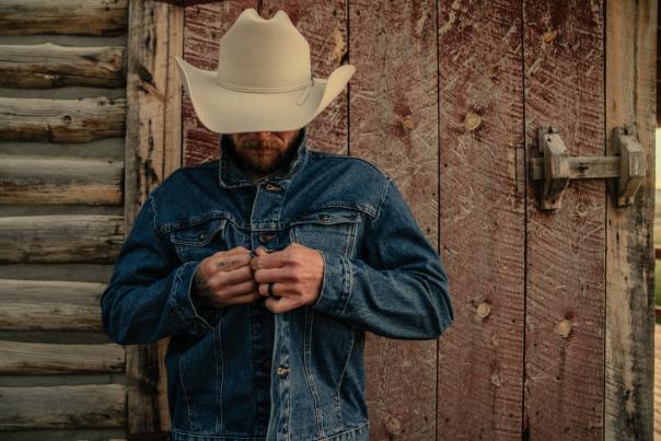 A cowboy wearing a white cowboy hat and denim jacket stands in front of a weathered wooden building near Buffalo, Wyoming. The rustic setting and traditional western attire reflect Wyoming's cowboy culture.