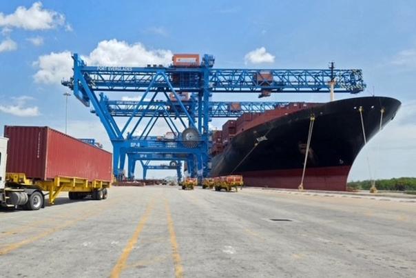 A cargo container truck drives past a black and red cargo ship at berth on a sunny day.