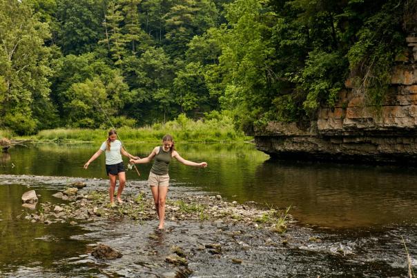 Two kids walking through water with fishing poles at a state park, with bluffs and trees behind them.