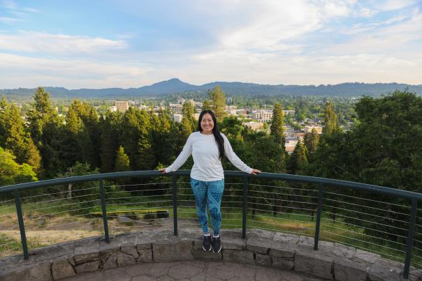 A woman stands by the railing at the top of Skinner Butte with the city view behind her.